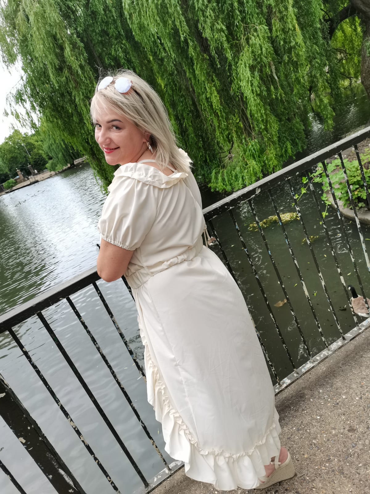 Kamilla Kostrzewa ubrana w białą, długą sukienkę. Stoi opierając się o barierkę, a przed nią jest rzeka, pływają w niej kaczki. Na brzegu rzeki drzewo. Kamila spogląda przez ramię i uśmiecha się.