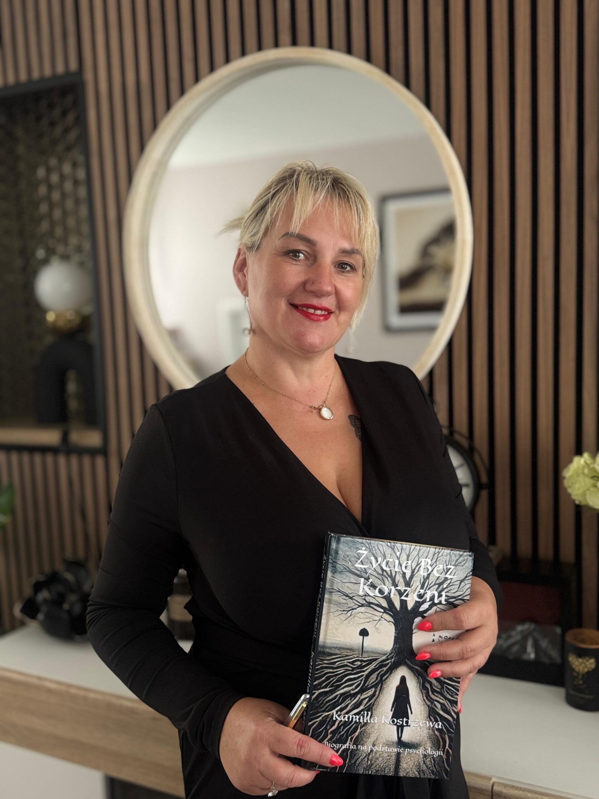 Kamilla Kostrzewa stoi i trzyma swoją książkę Życie bez korzeni. Jest ubrana w czarną sukienkę. Ma związane blond włosy, a na ustach czerwoną szminkę.