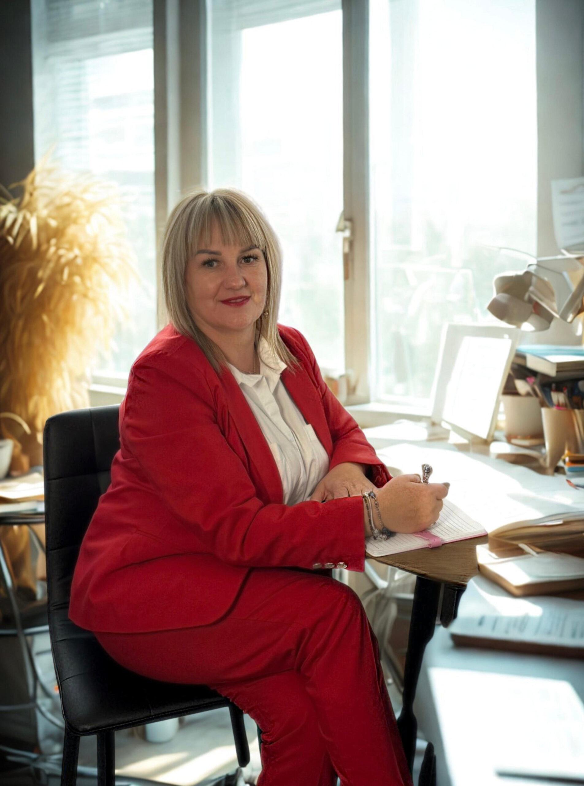 Kamilla Kostrzewa podczas pracy. Jest ubrana w czerwony garnitur. Siedzi przy biurku i pisze.